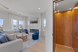 A modern living room with a sofa, armchair, coffee table, fireplace, and large windows. - NEW! Heated Pool, Elevator, 3min Beach Walk, Bunks, Quiet Family Retreat (Holden Beach)