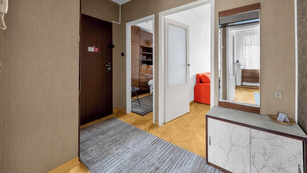 A hallway with a wooden floor, a grey rug, a wooden door, and a mirror reflecting a room with a red chair and a wooden floor. - Afrykańska Standard Apartment (Warszawa)