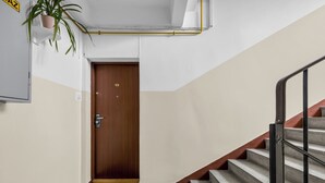 A hallway with a wooden door, a potted plant, a doormat, and a staircase with a metal railing. - Afrykańska Standard Apartment (Warszawa)