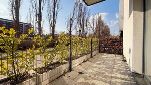 Una zona exterior empavedada amb una tanca de metall, arbres i un edifici al fons.