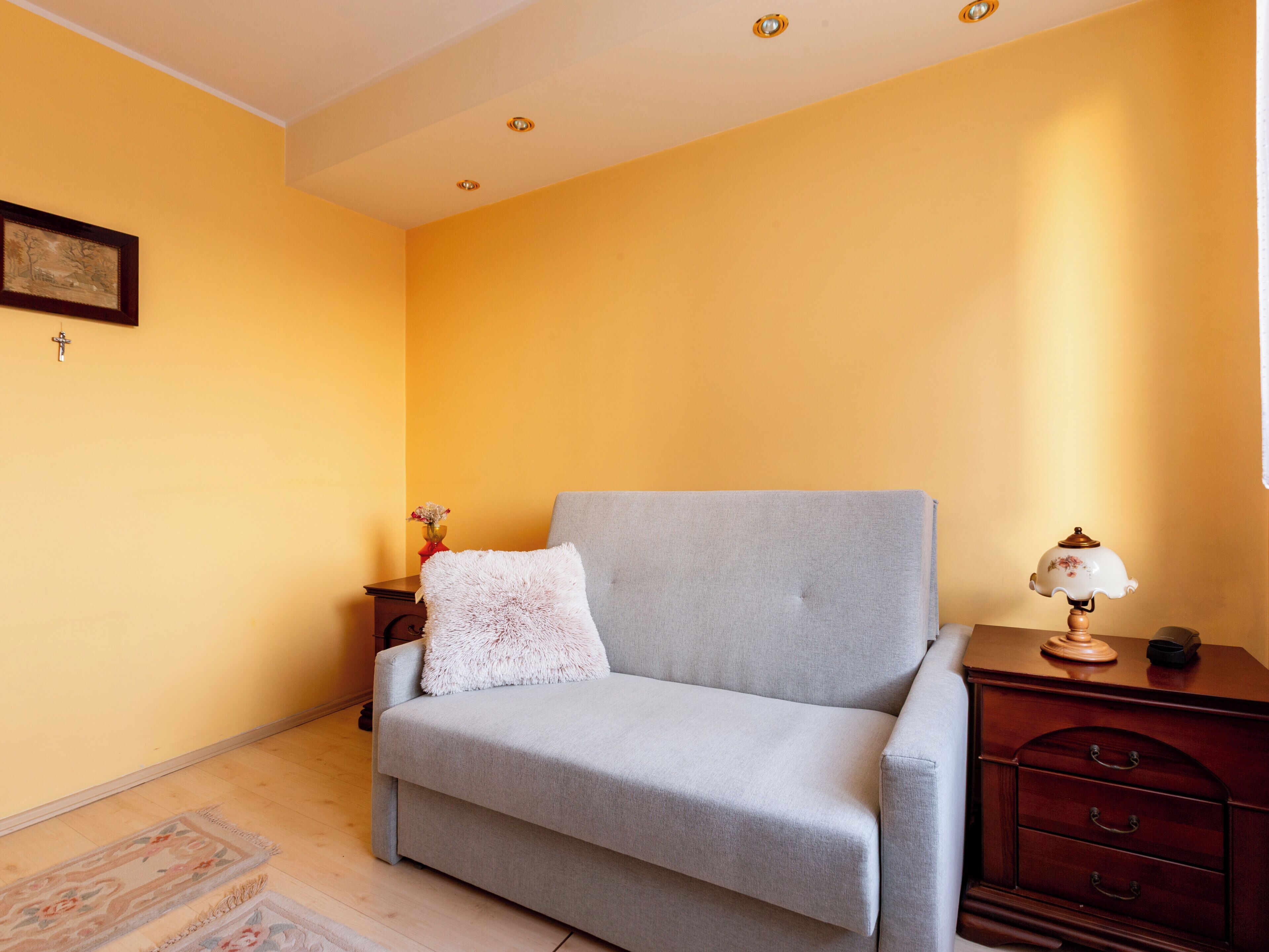 A room with a grey sofa, a wooden nightstand, a lamp, a framed picture, and a small table with a vase.