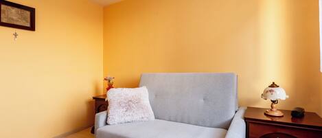 A room with a grey sofa, a wooden nightstand, a lamp, a framed picture, and a small table with a vase.