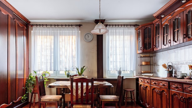Een traditionele keuken met houten kastjes, een eettafel met stoelen en een raam met dunne gordijnen.