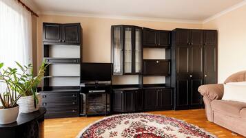 Un soggiorno con televisione, mobili contenitori in legno scuro, un divano beige e un tappeto decorato.