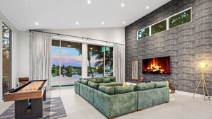 Ein modernes Wohnzimmer mit Billardtisch, einem grünen Sofa, einem Kamin und einem großen Fenster mit Blick auf einen Pool.