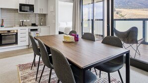 A modern kitchen with white cabinets, a microwave, and an oven. There is a dining area with a wooden table and chairs. - Mountain Ash View - Remarkable Apartment (Queenstown)
