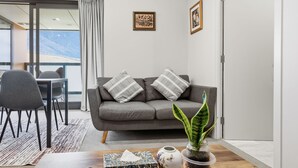 A modern living room with a grey sofa, two decorative pillows, a wooden coffee table, a remote control, a tissue box, a vase with a plant, and framed artwork on the wall. - Mountain Ash View - Remarkable Apartment (Queenstown)