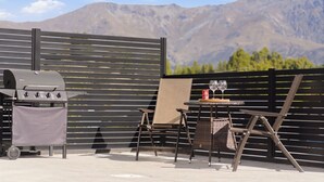 Une terrasse extérieure avec un barbecue, deux chaises, une table avec un plateau en verre et une vue sur les montagnes.