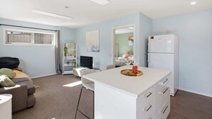 Un salon moderne avec un canapé, un coin repas avec une table blanche, une cuisine équipée d’un réfrigérateur et une fenêtre avec des rideaux.