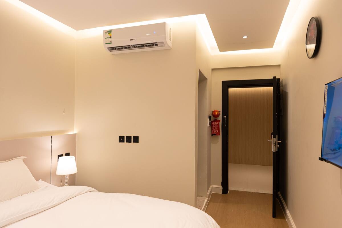 A hotel room with a bed, bedside lamp, air conditioning unit, and a door leading to another room.