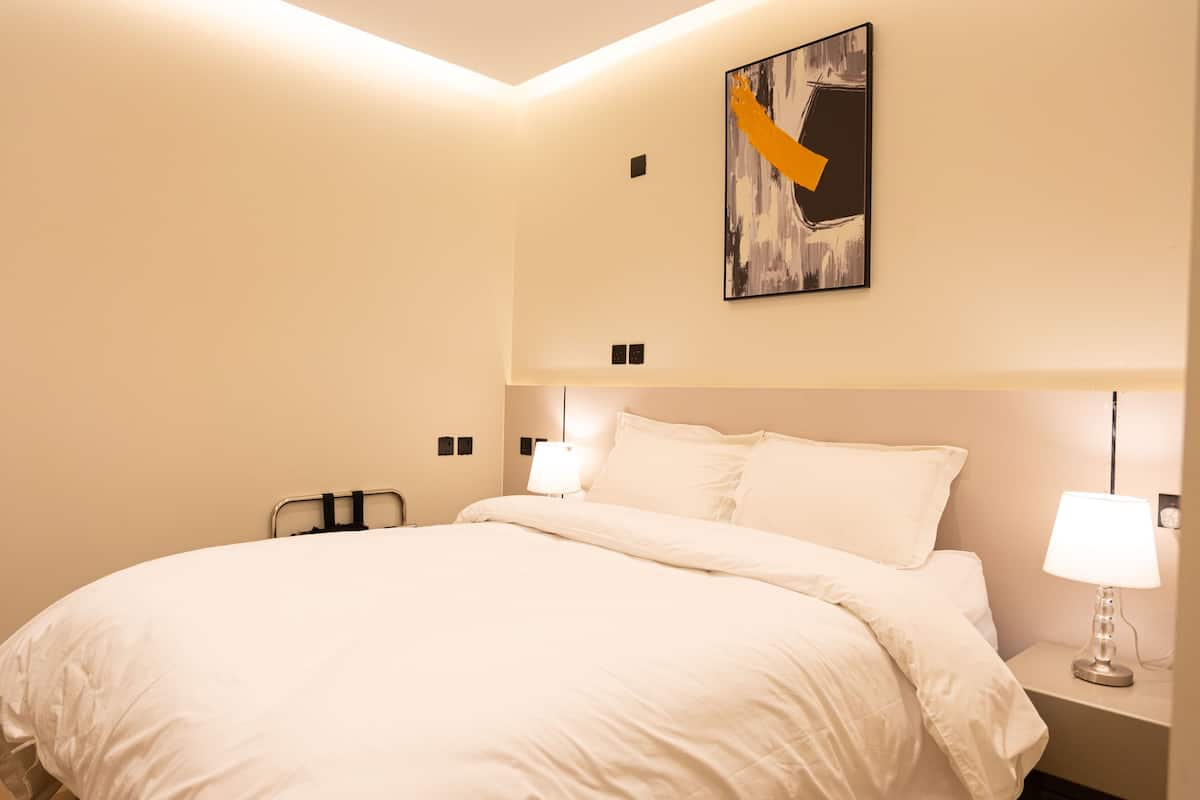 A hotel room with a large bed, a nightstand with a lamp, a wall-mounted artwork, and a ceiling light.