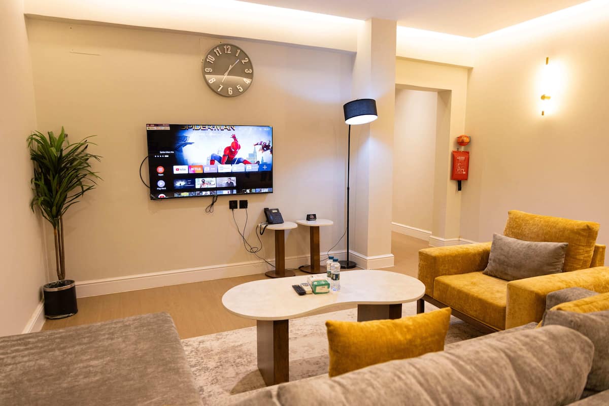 A living room with a flat-screen TV, a clock, a plant, a coffee table, and yellow sofas.