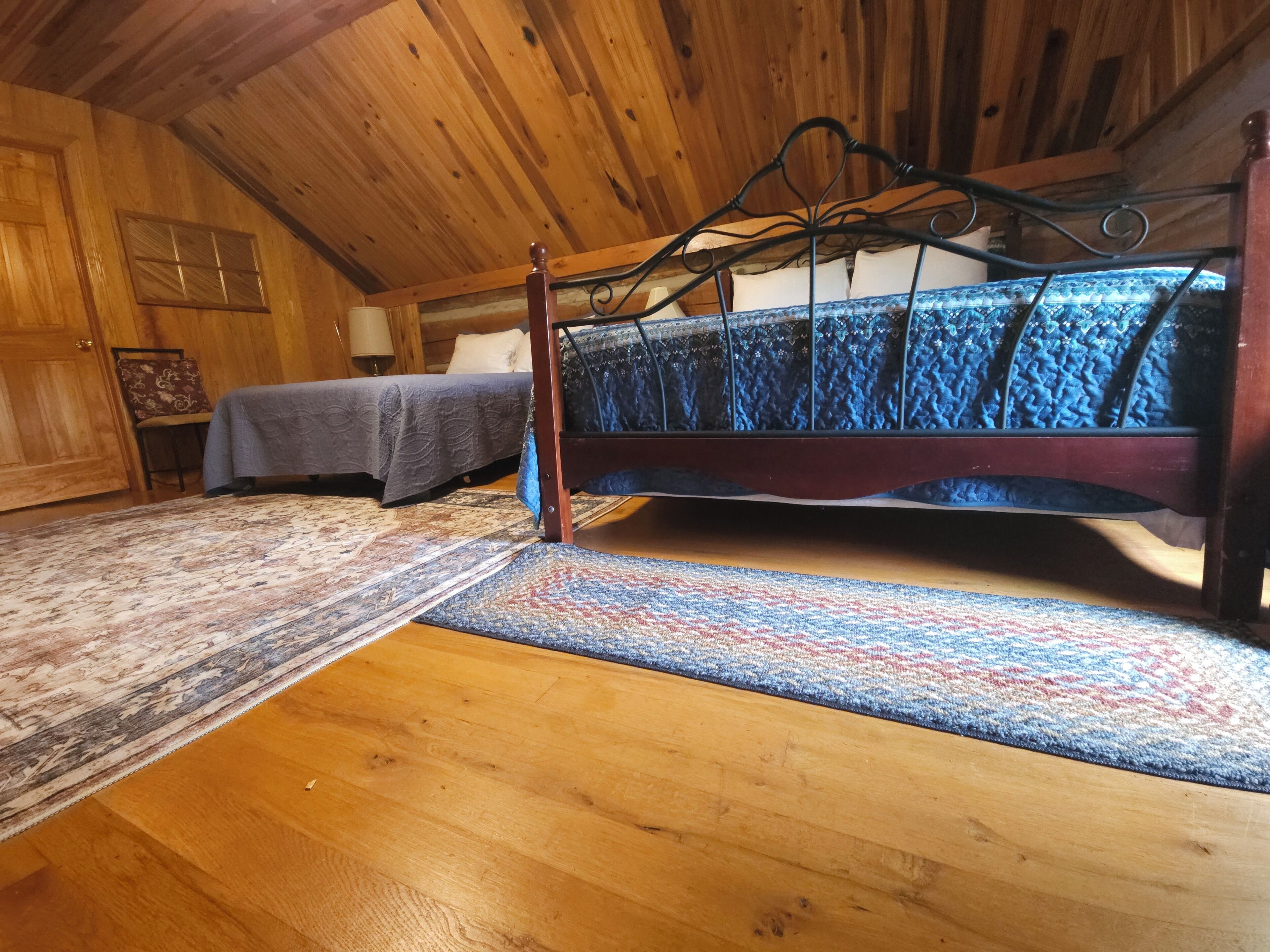 A wooden cabin interior with a bed, a chair, a rug, and a wooden door.