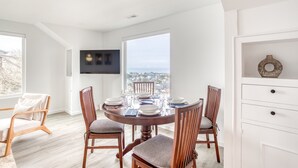 Uma área de refeições com mesa redonda posta para quatro pessoas, cadeiras de madeira, uma televisão fixada na parede e vista da cidade através de amplas janelas.