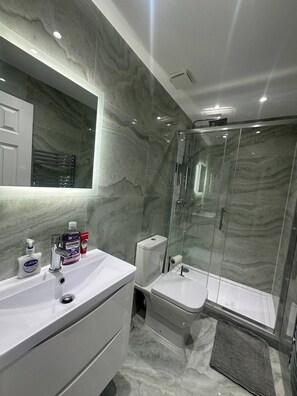 A modern bathroom with a marble wall, a glass shower enclosure, a white sink, and toiletries on the counter. - Luxury Apartment in Knightsbridge (London)