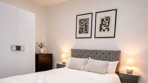 A neatly made bed with a white duvet and rolled towels, a tufted headboard, bedside tables with lamps, and a wall-mounted artwork. - Luxury Apartment in Knightsbridge (London)