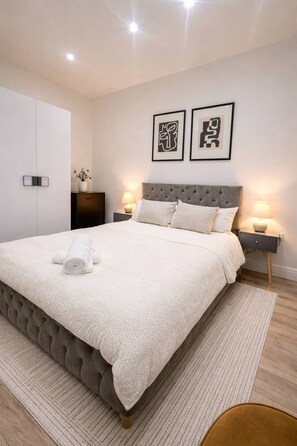 A neatly made bed with a white duvet and rolled towels, a tufted headboard, bedside tables with lamps, and a wall-mounted artwork. - Luxury Apartment in Knightsbridge (London)