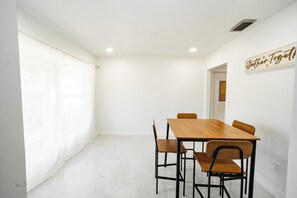 Ein Essbereich mit einem Holztisch und Stühlen, einer weißen Wand mit dem Schriftzug 'Gather Together' und einem Fenster mit durchscheinenden Vorhängen.