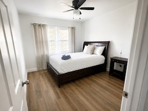 Ein Schlafzimmer mit einem Bett, einem Nachttisch, einem Deckenventilator und einem Fenster mit Vorhängen.