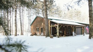 Una cabaña de madera rústica con un porche cubierto y una bandera estadounidense, rodeada de nieve y árboles.