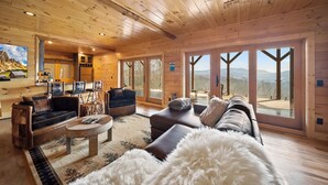 Ein Holzhütten-Interieur mit einer Couch, Sesseln, einem Couchtisch und großen Fenstern mit Blick auf die Berge.