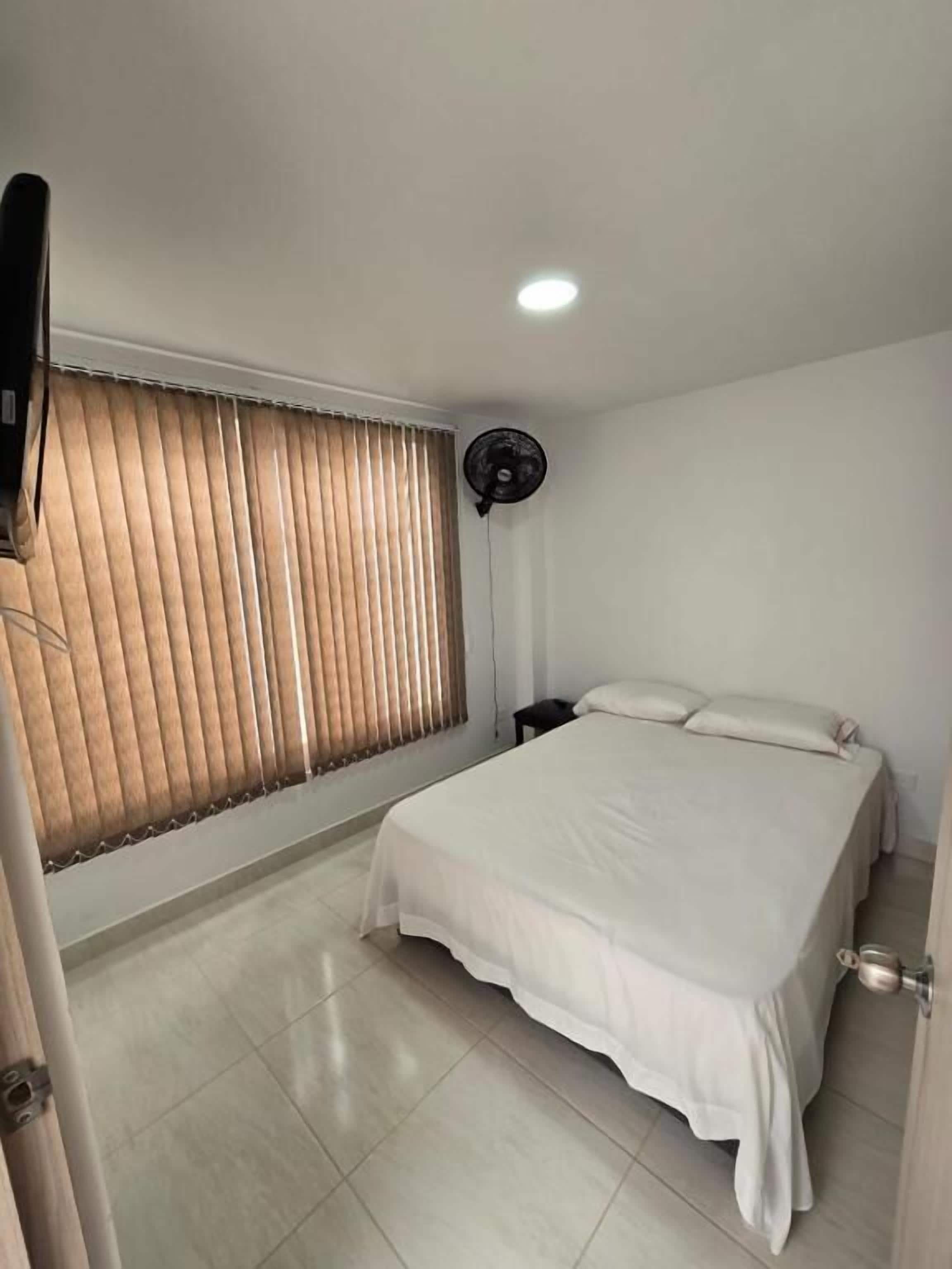 A bedroom with a bed, a standing fan, and a window with blinds.