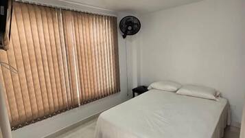 A bedroom with a bed, a standing fan, and a window with blinds.