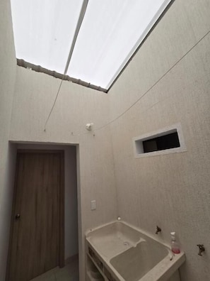 A bathroom with a skylight, a wooden door, a small window, and a sink with a soap dispenser.