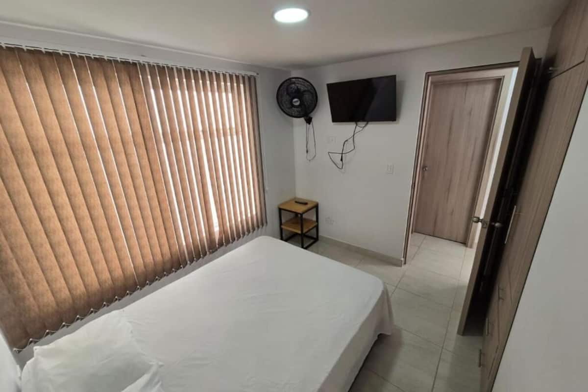 Une chambre à coucher avec un lit, un ventilateur sur pied, une télévision et une porte.
