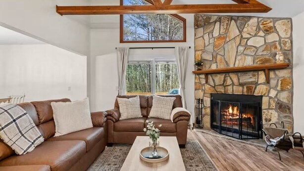 A living room with a stone fireplace, a brown leather sofa, a wooden coffee table, and a view of the outdoors through a large window. - Owl & Ember - Walk to Coosawatee Resort Center! Grill, 2 Kings, 3 Smart TV's (Ellijay)