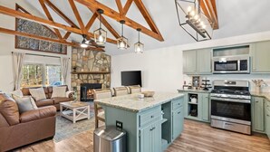 A spacious living room with a stone fireplace, a large brown leather sofa, and a kitchen with light blue cabinets and stainless steel appliances. - Owl & Ember - Walk to Coosawatee Resort Center! Grill, 2 Kings, 3 Smart TV's (Ellijay)