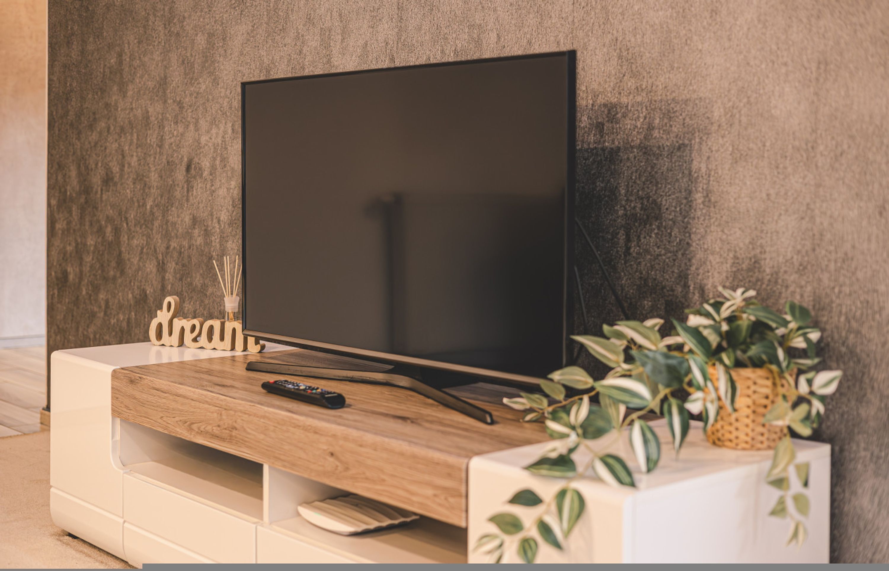 Ein Flachbildfernseher auf einer Holz-Unterhaltungseinrichtung mit Fernbedienung, einer kleinen Pflanze und dem dekorativen Wort 'dream' auf der linken Seite.