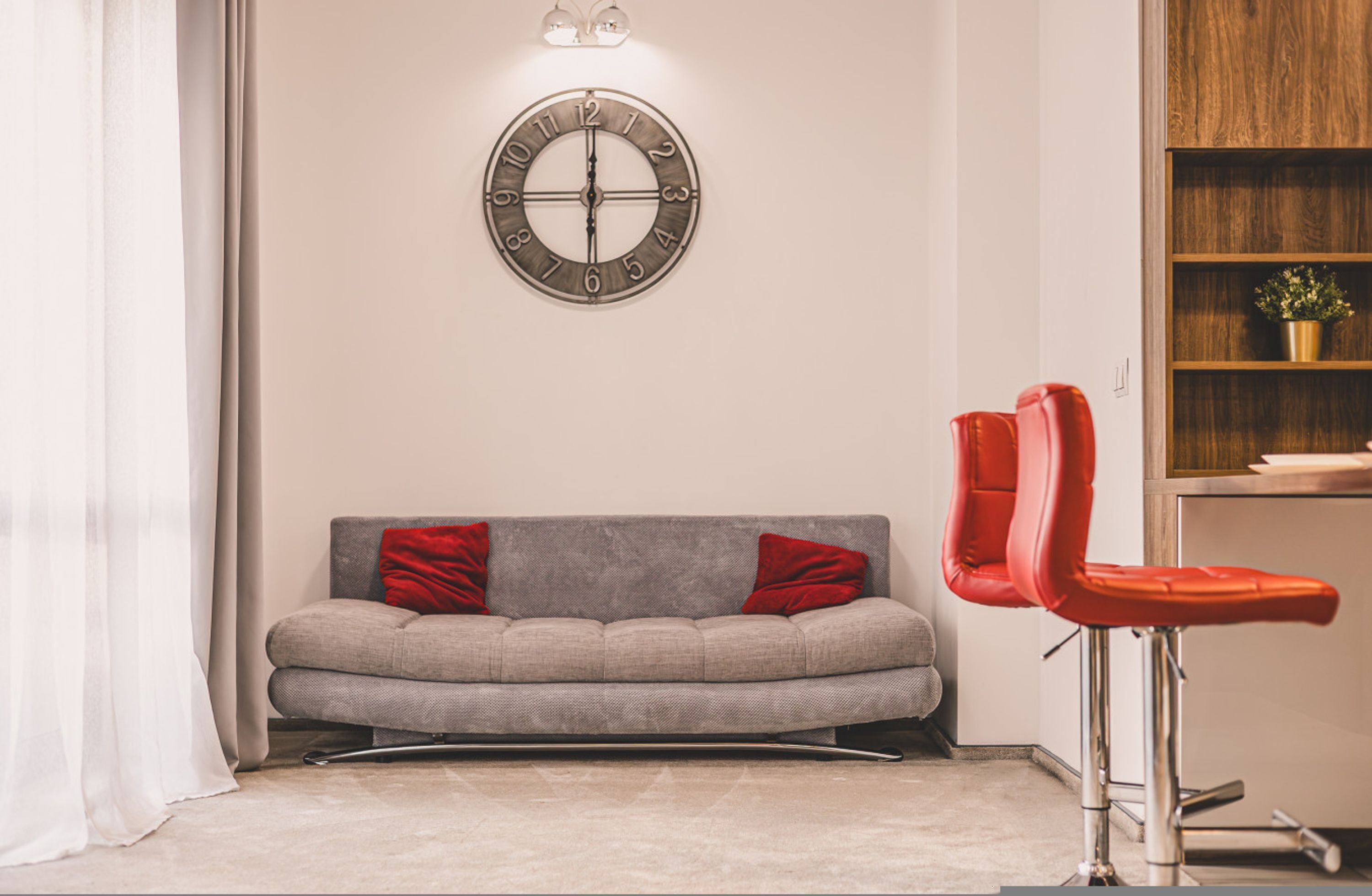 A modern living room with a grey sofa, red cushions, a round wall clock, and a red bar stool.