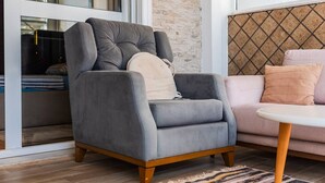 Un fauteuil gris aux pieds en bois, un canapé rose et un tapis à motifs dans une pièce dont un mur est en briques et qui possède une fenêtre.