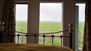 A room with a bed, a view of a green field through large windows, and a wooden ceiling.