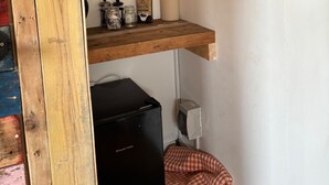A kitchen area with a wooden ceiling, a refrigerator, a shelf with jars, a framed picture, and a wall-mounted electrical box.