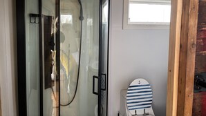 A bathroom with a glass shower enclosure, a toilet with a striped seat, and a window above the toilet.