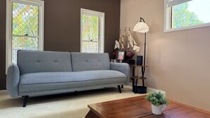 Un soggiorno con un divano grigio, un tavolino in legno e una lampada da terra. Sono presenti due finestre con vetrate decorative.