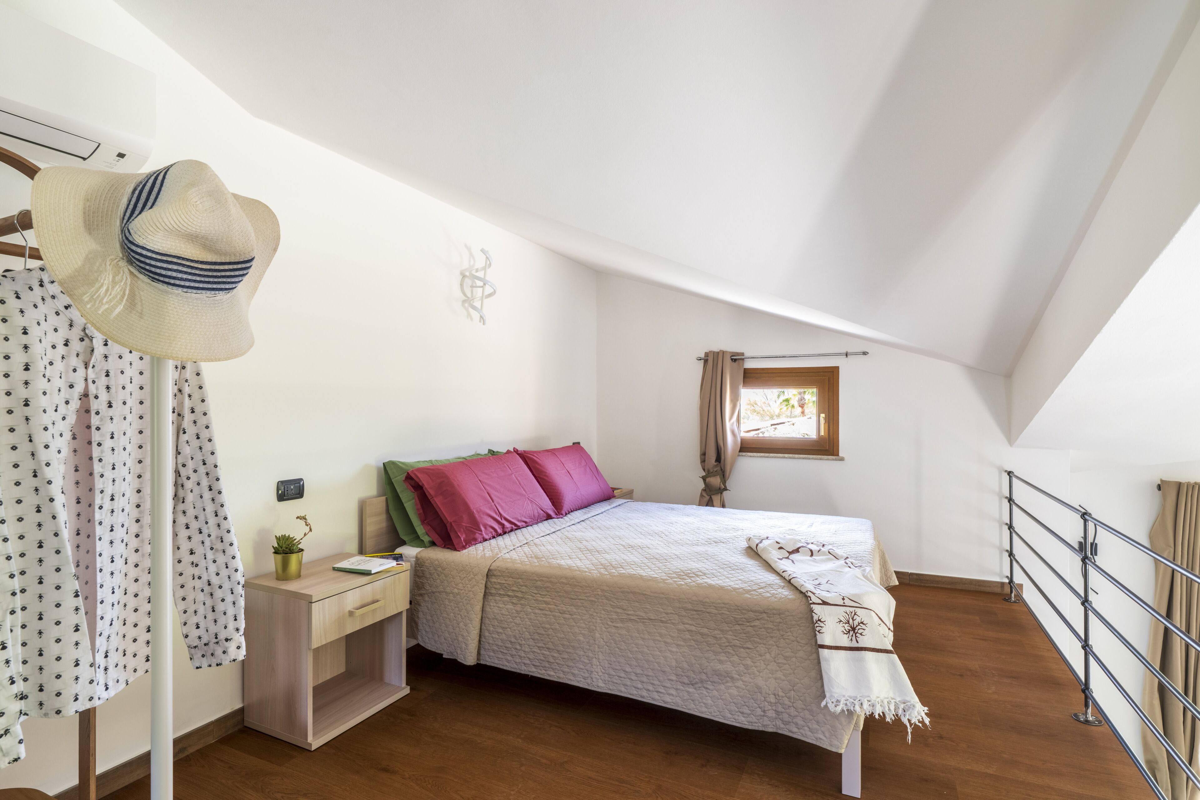 A bedroom with a bed, a nightstand, a window, and a hat hanging on a wall.
