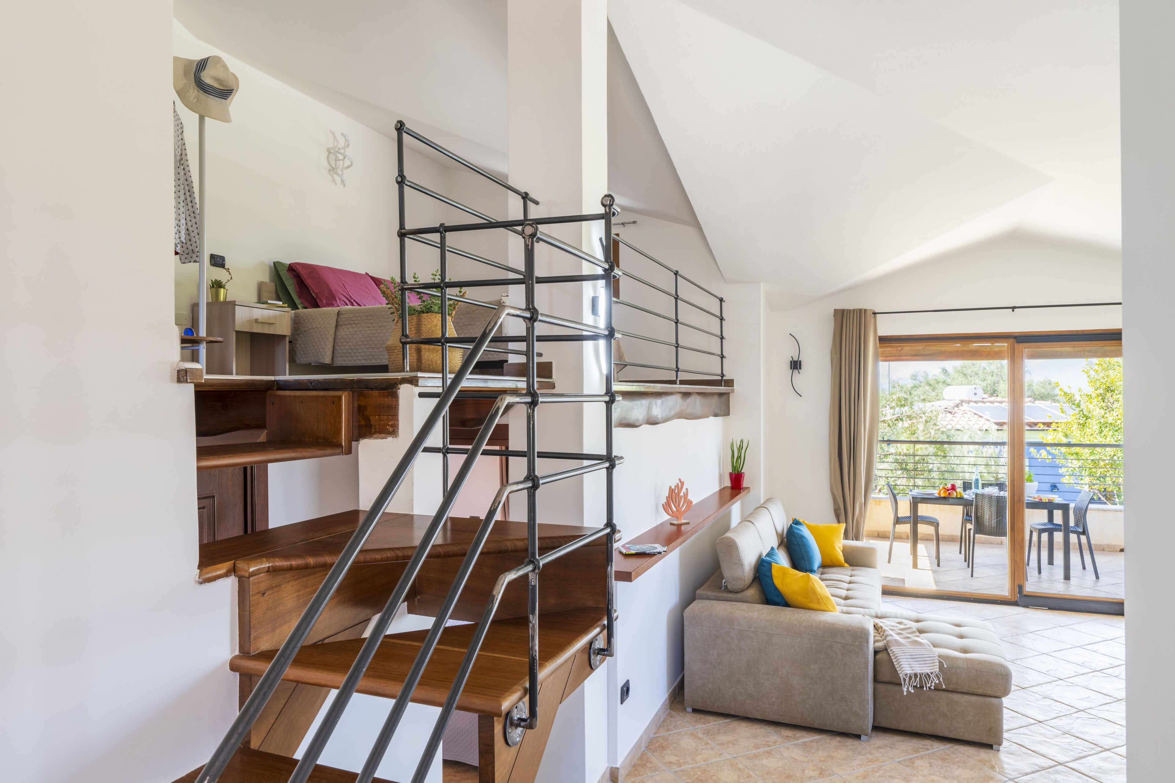 Een modern interieur met een houten trap, een bank met kleurrijke kussens en een balkon met tuinmeubilair.