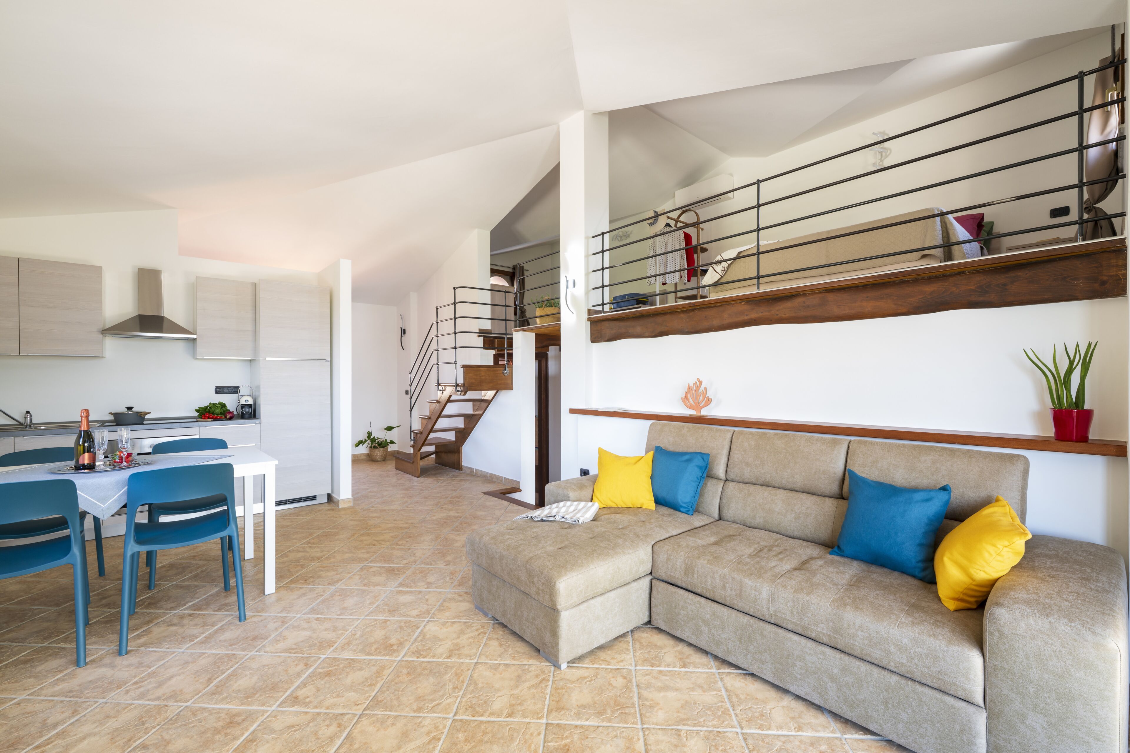 Une cuisine moderne avec des meubles blancs, un coin repas avec des chaises bleues, un canapé beige agrémenté de coussins colorés et un escalier menant à un étage supérieur.