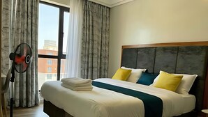 A neatly made bed with a wooden headboard, a standing fan, and a view of buildings through the window. - Luxury 6-Bed Penthouse with Sky Terrace (Nairobi)