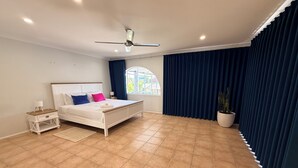 Ein Schlafzimmer mit einem Bett, einem Nachttisch, einem Deckenventilator, einem Fenster mit Vorhängen und einer Topfpflanze.