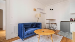 Ein Zimmer mit einem blauen Sofa, einem runden Holztisch, einer Lampe, einer kleinen Küchenzeile mit Kühlschrank und einem gemusterten Teppich.