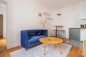 A room with a blue sofa, a round wooden table, a lamp, a small kitchen area with a fridge, and a patterned rug. - Blueground | Europe, elevator, nr Parc Monceau (Paris)