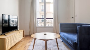 Ein Zimmer mit einem HolztV-Schrank, einem runden Tisch, einem blauen Sofa und einem Fenster mit Balkonblick.