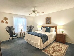A bedroom with a bed, two chairs, a ceiling fan, a nightstand with a lamp, a framed picture, and a patterned rug.
