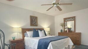 Quarto com cama, duas mesinhas de cabeceira, uma cadeira, cômoda, ventoinha no teto e espelho.