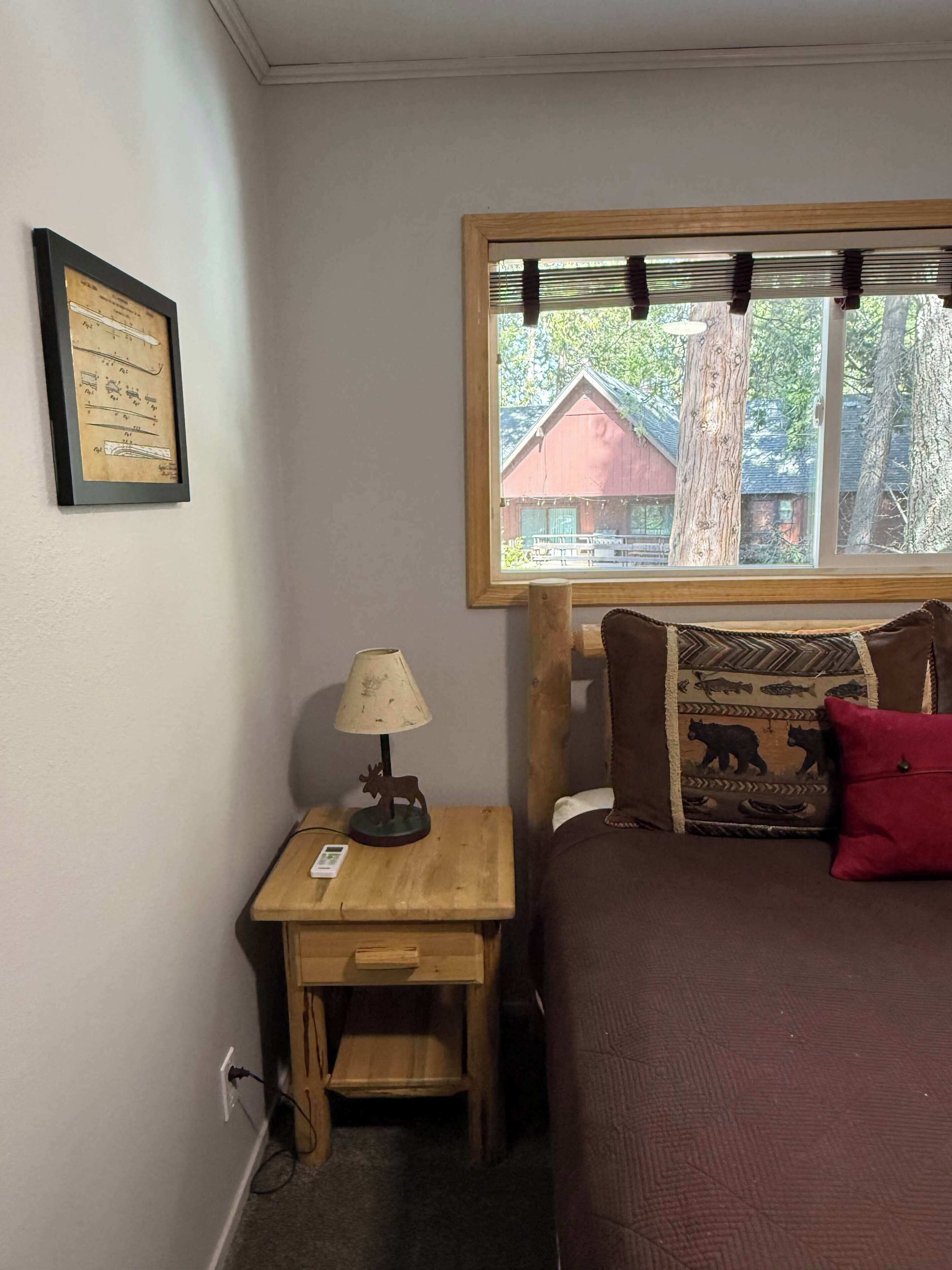 Une chambre à coucher avec un lit, une table de chevet avec une lampe, un certificat encadré accroché au mur et une fenêtre donnant sur un bâtiment rouge et des arbres.