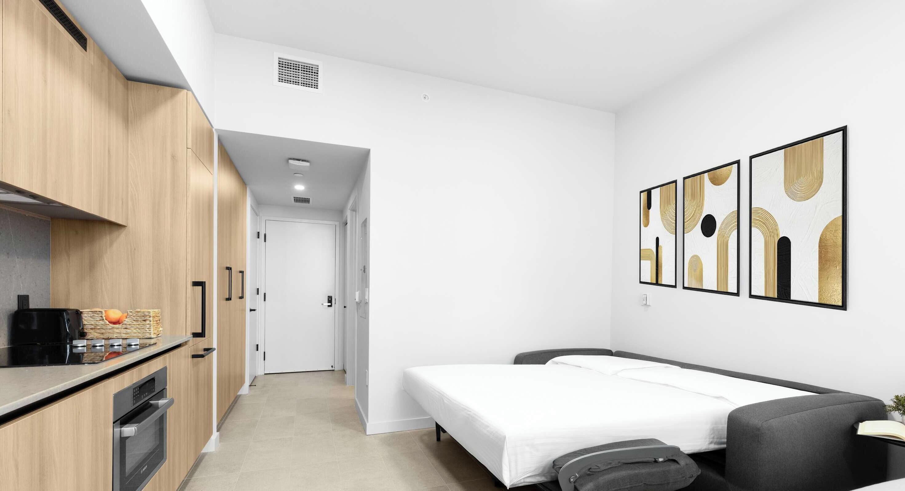 A compact hotel room with a kitchenette, a bed, a bedside table, and artwork on the wall.